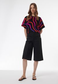 Camisa negra de manga corta con vibrantes patrones de olas en púrpura, rojo y rosa; combinada con pantalones cortos negros hasta la rodilla y sandalias negras con tiras.