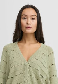 Hellgrüne gestrickte Strickjacke mit V-Ausschnitt, Knopfleiste und offenem Muster mit horizontalen Streifen und kleinen Ausschnitten.