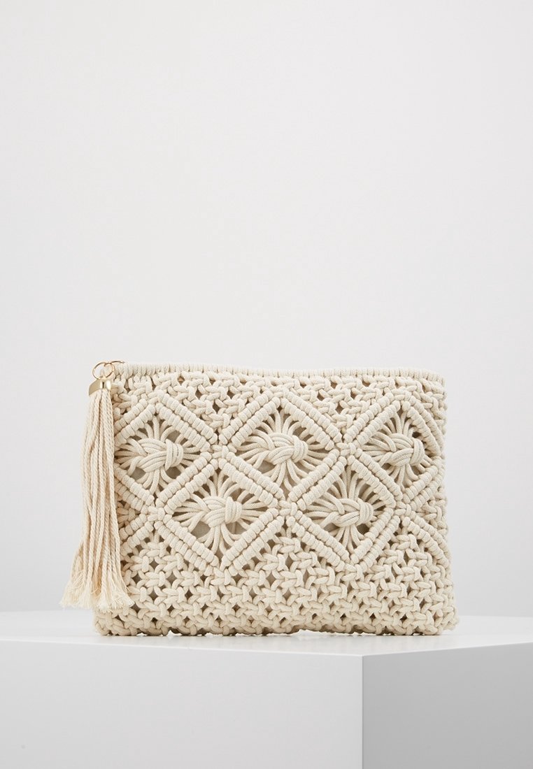 Pochette rectangulaire en macramé couleur crème avec motif de losanges et nœuds, ornée d'un pompon sur la fermeture éclair, posée sur un socle blanc.