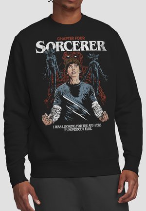 Person wearing black sweatshirt with graphic of a tense boy surrounded by shadowy creatures and text "Chapter Four Sorcerer" and a quote below.