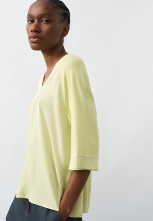 Junge Person mit geflochtenem Haar, die ein lockeres hellgelbes V-Ausschnitt-Oberteil und dunkle Hosen trägt, steht vor einem schlichten hellen Hintergrund.