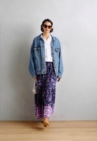 Veste en denim sur une chemise blanche, associée à une jupe maxi à motifs violets. Les accessoires comprennent des lunettes de soleil surdimensionnées et un petit sac blanc.