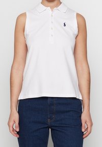 Bílé bezrukávové polo tričko s knoflíkovou légou, límečkem a malým modrým vyšitým logem na levé hrudi.