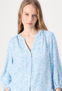 Blusa azzurro chiaro con bottoni, motivo floreale bianco, scollatura rotonda e maniche lunghe con polsini elastici, realizzata in un tessuto morbido.