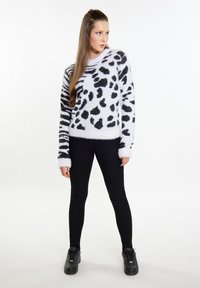 Pluizig wit sweater met zwart dierenprint, ronde halslijn en lange mouwen, gecombineerd met zwarte skinny broeken en zwarte sneakers.