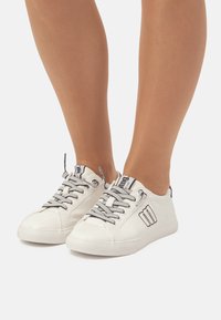 Zapatillas blancas de material suave, con cordones grises, puntera redonda y un detalle del logo en el costado. Suela plana y diseño de caña baja.