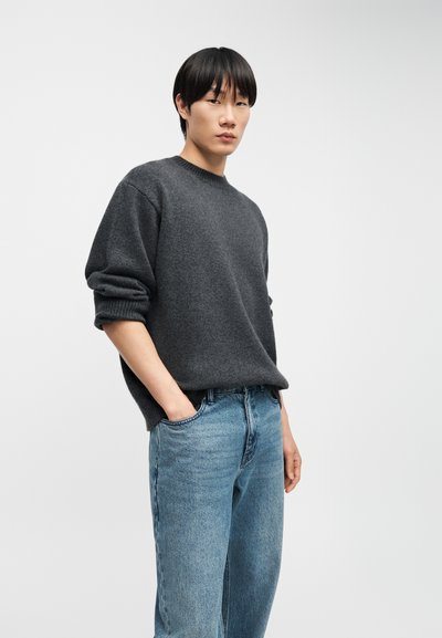 Jeune homme aux cheveux noirs portant un pull gris foncé et un jean bleu, debout avec une main dans la poche devant un fond clair uni.