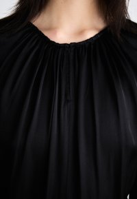 Musta paita, jossa on kerätty kaula-auko, napitusyksityiskohtia ja sileä kangas. Suunnittelussa on pehmeitä laskoksia, jotka ulottuvat kauluksesta.