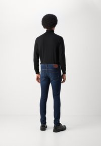 Top de manga larga negro, ajustado, combinado con jeans ajustados azul oscuro con un parche de cuero marrón y bolsillos traseros. Zapatos negros completan el look.