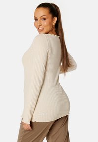 Beige geribbelde lange mouw top met een scalloped halslijn, strak ontwerp en een licht uitlopende zoom; gecombineerd met een bruine broek.