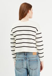 Suéter cropped blanco con rayas negras, con mangas largas y escote redondo. Combinado con jeans azul de tiro medio y una etiqueta de marca en tono marrón.