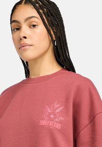Vaaleanpunainen collegepaita, jossa on pyöreä kaula-aukko. Paidassa on kukkakuosi ja rinnassa valkoisella painettuna "TIMBERLAND EST. 1973". Pehmeä rakenne, rento istuvuus.