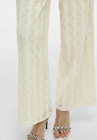 Pantalones de pierna ancha en una tela de punto color crema, con un patrón texturizado en forma de olas. Terminado con un dobladillo que cae suavemente. Combinados con sandalias acentuadas en plata.