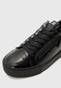 Sneakers neri con finitura lucida in vernice, dotati di punta rotonda, suola in gomma testurizzata e lacci neri. Include dettaglio con cerniera laterale.