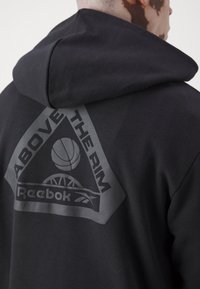 Sweatshirt à capuche noire avec un grand motif de triangle gris au dos, accompagné du texte "ABOVE THE RIM" et d'un graphique de basketball.