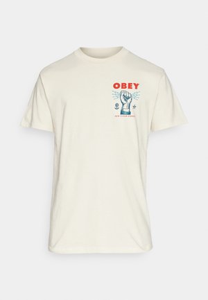 T-shirt blanc cassé avec texte rouge "OBEY", graphique d'un poing bleu serré, et petits symboles de fleur et d'étoile sur la poitrine.