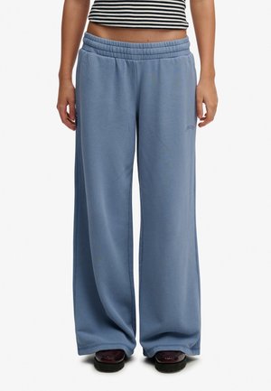 Persona que lleva pantalones de chándal azules y sueltos y zapatos oscuros, de pie con las manos relajadas a los lados, recortada por debajo del pecho.