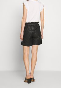 Black leather shorts with elastic waistband, paired with a white top featuring ruffled sleeves. Model shown from the back, wearing black sandals.