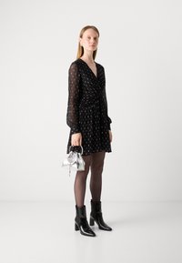 Abito nero con lunghe maniche trasparenti, design avvolgente e motivo a pois bianchi. Abbinato a una borsa argento e stivaletti neri. Tessuto morbido.
