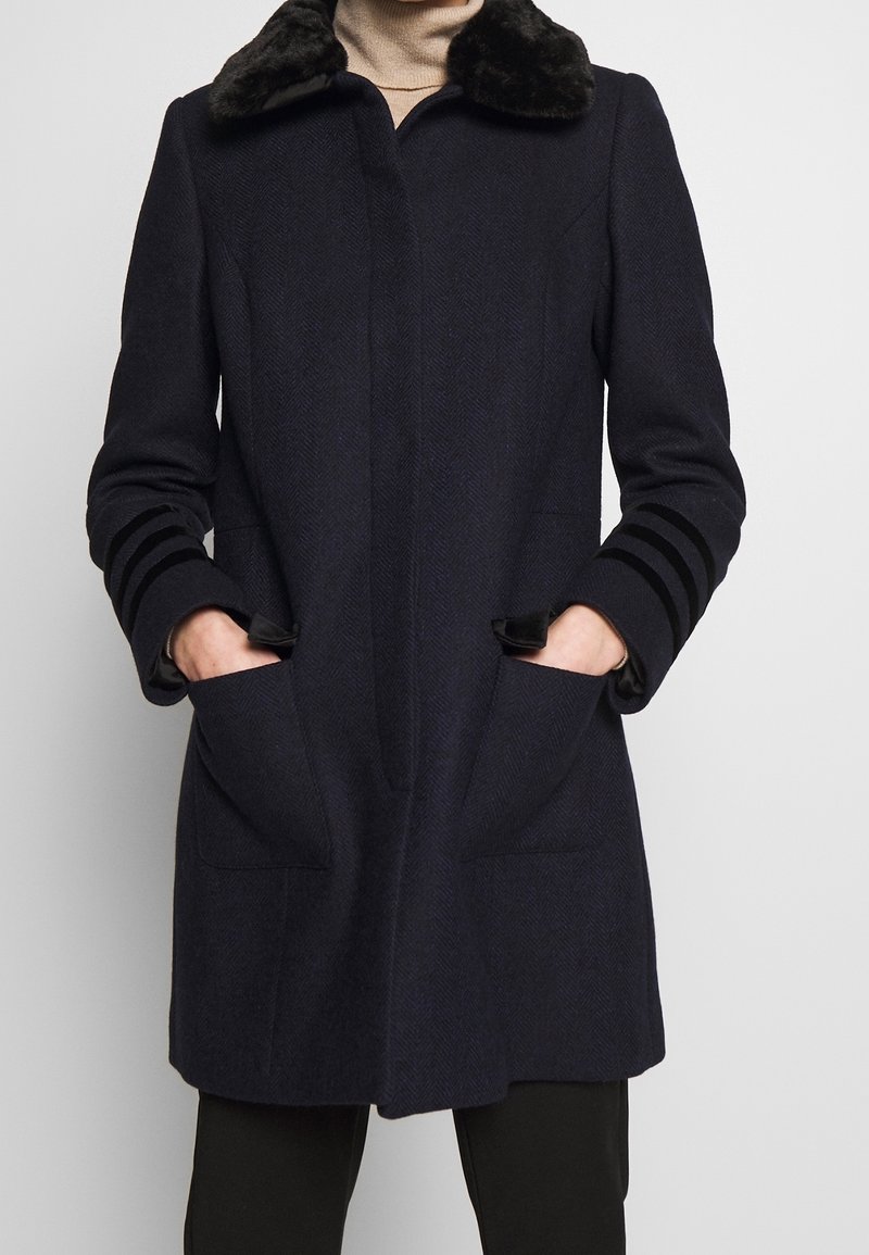 Personne portant un manteau bleu marine foncé avec un col en fourrure noire, des poches avant et trois bandes noires sur chaque manche, les mains dans les poches.