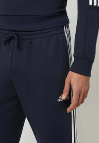 Pantaloni da jogging athletic blu navy con strisce bianche sui lati, vita con cordino e texture morbida. Presenta il logo Adidas sulla coscia.