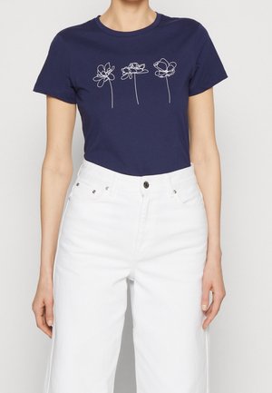 Personne portant un t-shirt bleu marine avec trois dessins de fleurs en ligne continue blanche et un pantalon blanc taille haute.