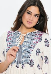 Broderet bluse med en cremefarvet bund, der har blå og pink blomster mønstre, en nøglehulhalsudskæring og en bindedetail ved kraven.
