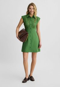 Abito verde corto con un corpetto in pizzo, maniche a cappuccio e chiusura a bottoni. Abbinato a una pochette marrone con dettagli in catena d'oro e scarpe slip-on.