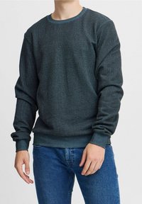 Pullover lavorato a maglia verde scuro con scollatura tonda, polsini e orlo a costine, texture liscia, indossato con jeans blu.