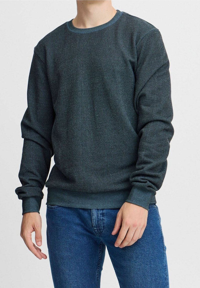 Pullover lavorato a maglia verde scuro con scollatura tonda, polsini e orlo a costine, texture liscia, indossato con jeans blu.