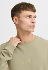 Sudadera verde claro con cuello y puños de canalé. Presenta una textura suave y un logo bordado en el centro. La modelo sostiene el cuello.