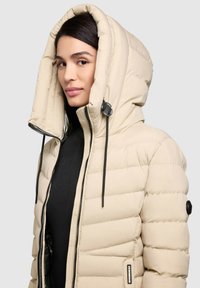 Bézs színű pehelykabát nagy, állítható kapucnival, függőleges steppelt mintázattal, elöl cipzárral és fekete húzózsinór részletekkel.