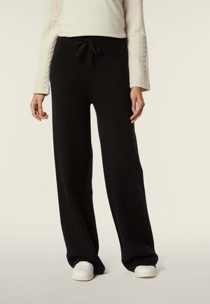 Schwarze, weit geschnittene Hose aus weichem Material, mit verstellbarem Kordelzugbund, kombiniert mit weißen Sneakers. Schlichtes Design, geeignet für die Freizeit.