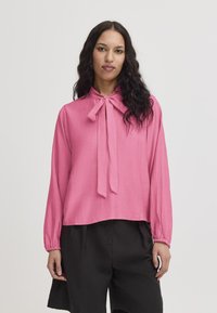 Blusa rosa con collo annodato, maniche lunghe e tessuto testurizzato. Abbinata a pantaloni neri, con una vestibilità comoda.