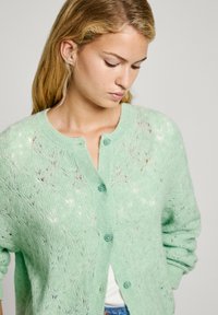 Minta zaļš adīts kardigans ar mežģīņu rakstu, apaļu kaklu un sešiem pogu aizdari priekšā. Garas piedurknes ar ribinātiem aproces galiem.