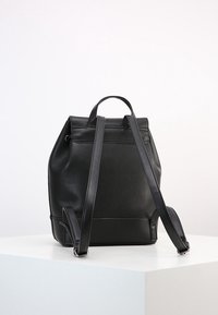 Sac à dos en cuir noir avec bretelles réglables et une poignée supérieure, présenté sur un socle blanc contre un fond uni.