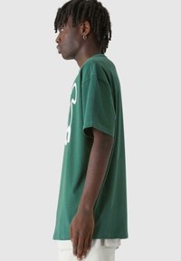 Žalias medvilninis T-shirt su balta grafika, trumpomis rankovėmis ir laisvu siluetu, derinamas su šviesiomis kelnėmis.