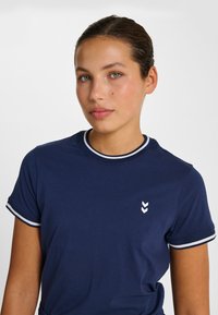 Mörkblå t-shirt med korta ärmar, vit kant och en liten vit chevron-logotyp på bröstet. Bomullsmaterial, slät textur.