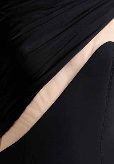 Juoda maudymosi apranga su klostuota tekstūra viršuje, kietas juodas audinys apačioje, o nude spalvos pamušalas matomas krašte, su lygiais siūlais.