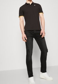 Czarna koszulka polo z żółto-niebieskim paskiem przy kołnierzyku, zestawiona z czarnymi, dopasowanymi dżinsami z efektami przetarcia. Całość dopełniają białe trampki.