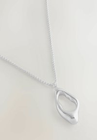 Srebrny naszyjnik z łańcuszka z abstrakcyjnym, pustym, organicznym zawieszeniem na jasnym tle.