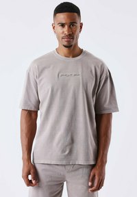 Camiseta de algodón gris claro con mangas cortas, que presenta un logo rectangular texturizado en el centro. Cuello redondo estándar y corte relajado.