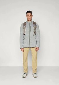 Gray fleece jacket with a zip closure, worn over tan pants. The individual also has a beige backpack with water bottles attached.