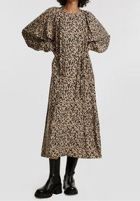 Vestido marrón con estampado abstracto negro, mangas globo y cintura ceñida con cinturón. Combinado con botas negras de tobillo gruesas. Diseño largo y fluido.