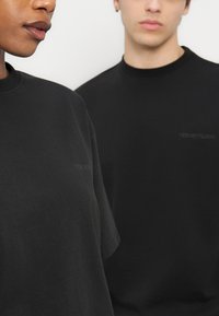 Δύο άτομα που φορούν απλά μαύρα μπλουζάκια με λαιμόκοψη, με διακριτικό κείμενο "YOUR TURN" στο στήθος, σε ουδέτερο φόντο.