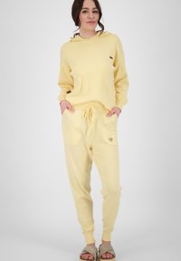 Geel hoodie en sweatpants set gemaakt van zachte stof. De hoodie heeft een voorzak, en de broek heeft twee grote zakken en een hartlogo.
