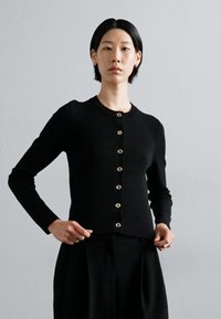 Svart strikket cardigan med rund hals og gullknapper foran. Fittet design med lange ermer og ribbestrikkede detaljer.