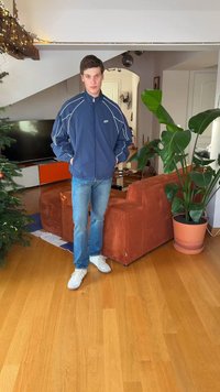 Plava sportska jakna s svijetlim prugama, plave traperice i bijele tenisice. Enterijer s drvenim podom, smeđom sofom i lončanicom.