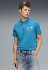 Turkizna polo majica s ovratnikom, kratkimi rokavi in belim logotipom BMW Motorsport; v kombinaciji s svetlo modrimi unikatnimi džinsami.