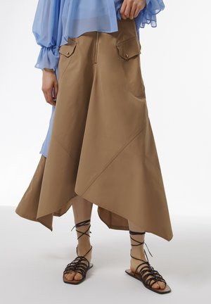 Asymmetrische bruine rok met twee voorkruiszakken, gemaakt van een zachte stof; gecombineerd met zwarte sandaaltjes met bandjes.